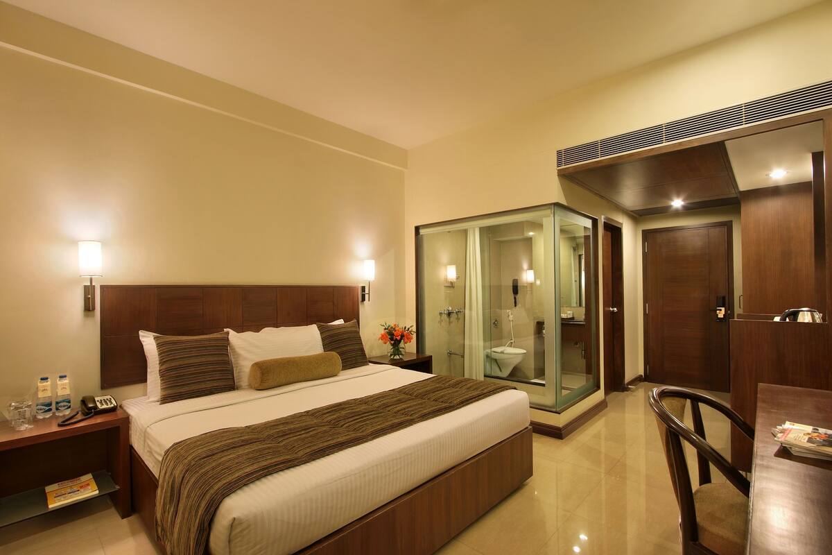 A hotel room with a large bed, bedside tables, a glass-enclosed bathroom, a wooden desk, and a chair.