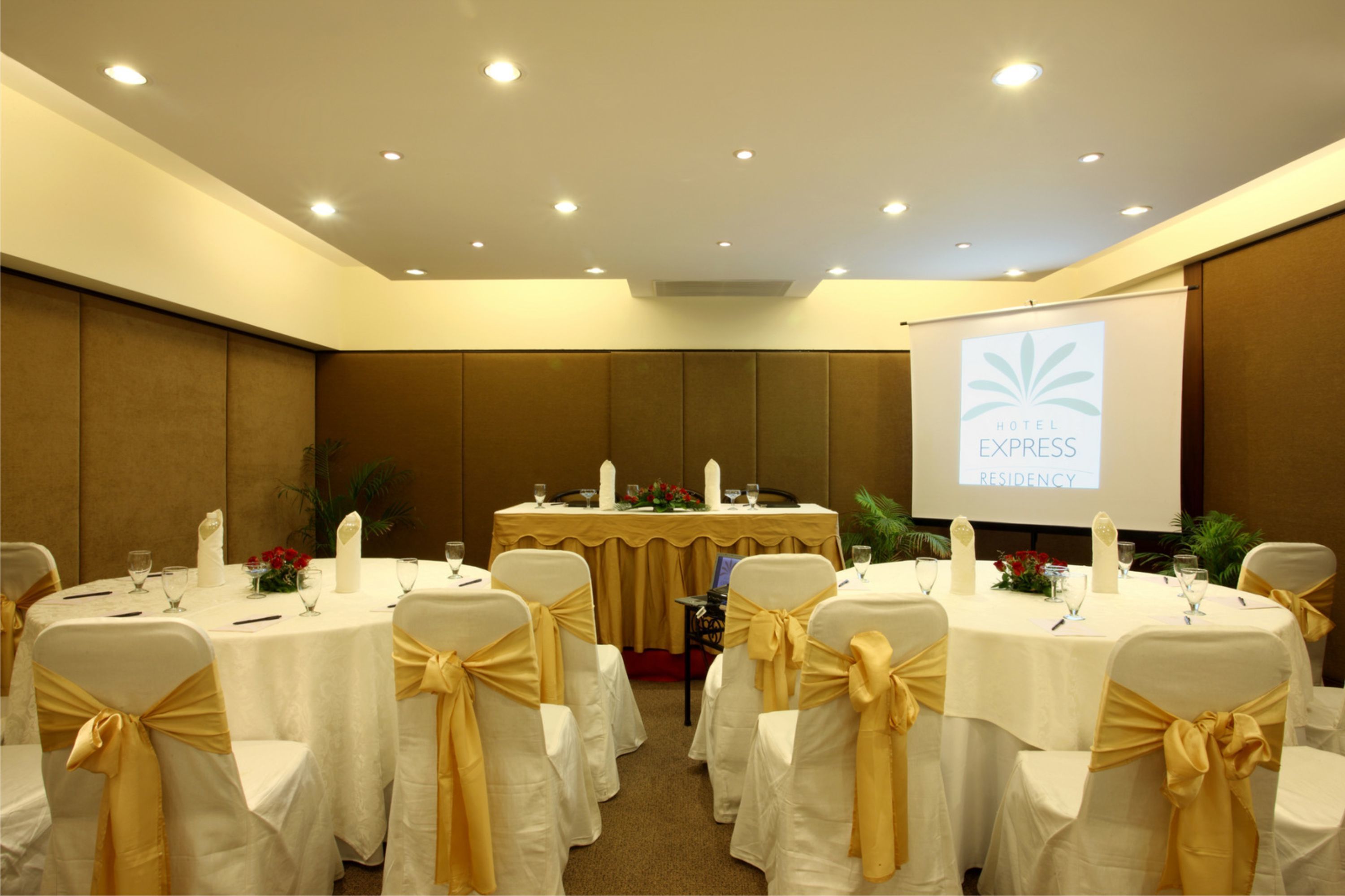 Un salón de conferencias con mesas redondas, manteles blancos y sillas con cintas doradas. Una pantalla proyecta 