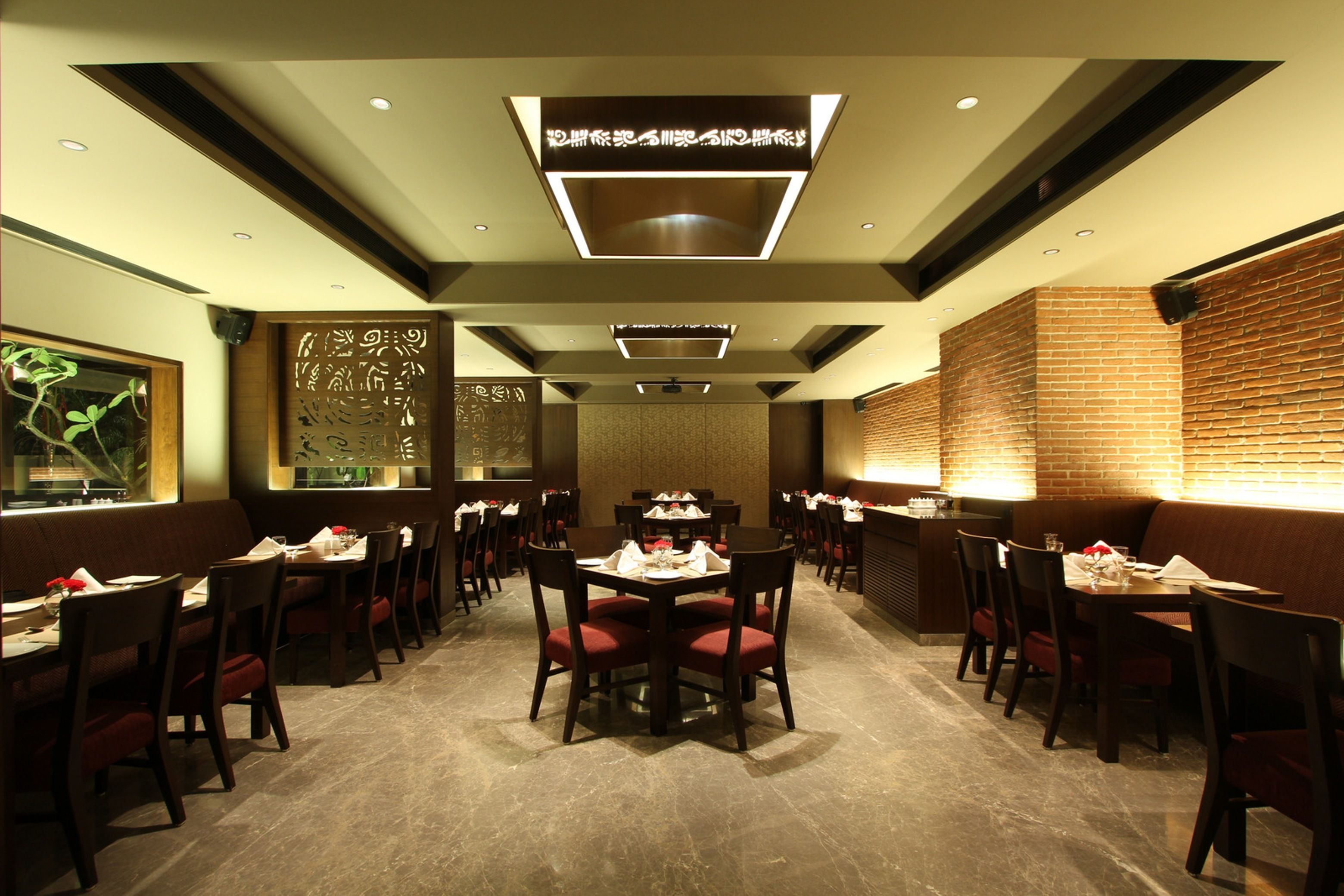 A restaurant with a modern interior, featuring a central dining area with round tables and red cushioned chairs, surrounded by brick walls and recessed lighting.