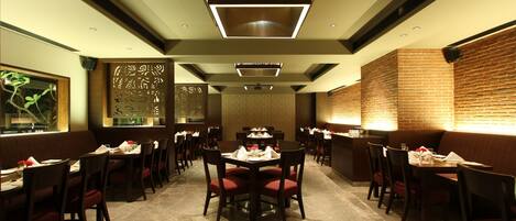 A restaurant with a modern interior, featuring a central dining area with round tables and red cushioned chairs, surrounded by brick walls and recessed lighting.