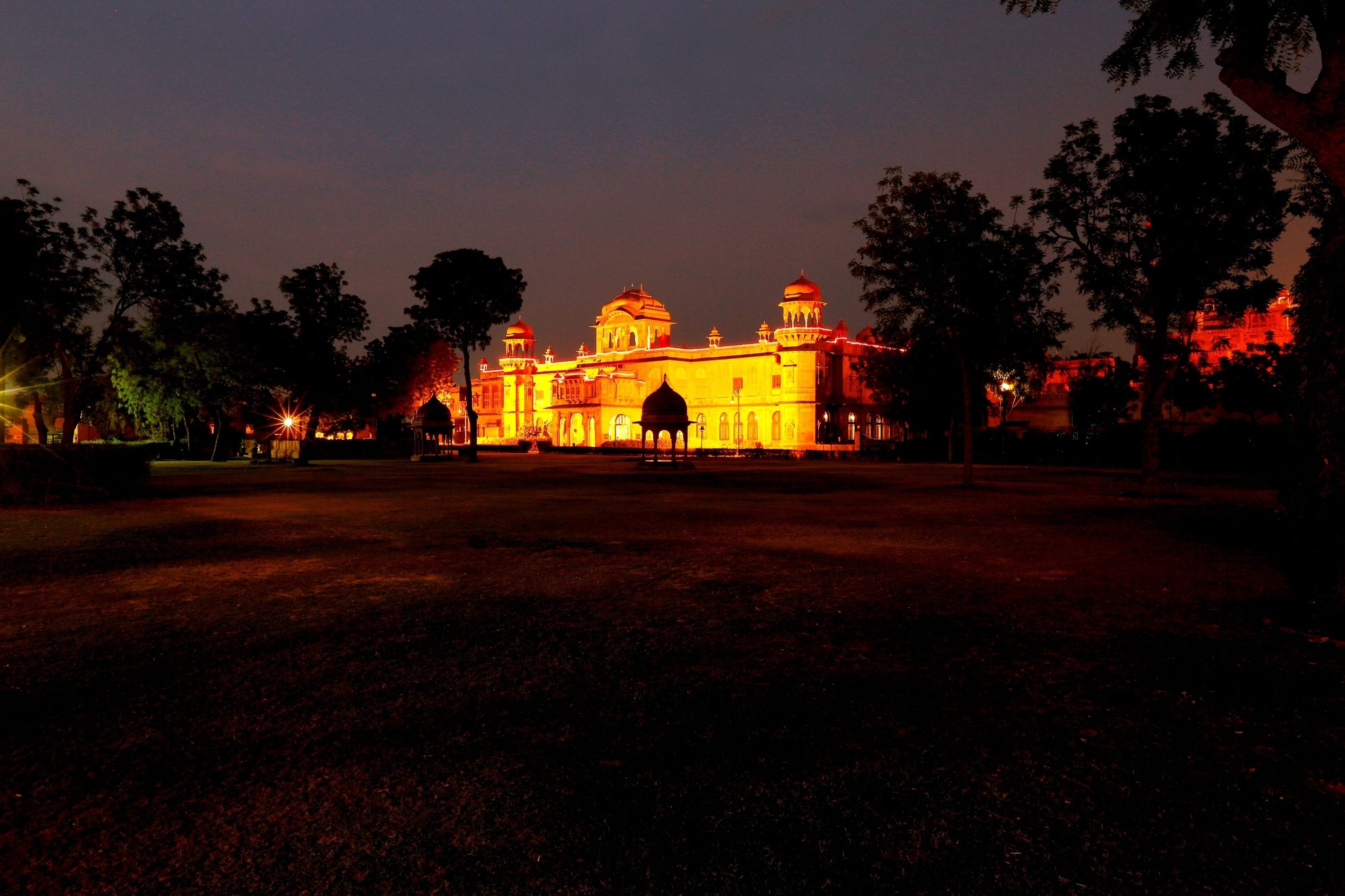 A large, illuminated building with multiple domes and arches, surrounded by trees and a paved area.