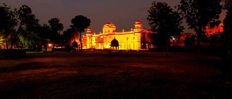 A large, illuminated building with multiple domes and arches, surrounded by trees and a paved area.