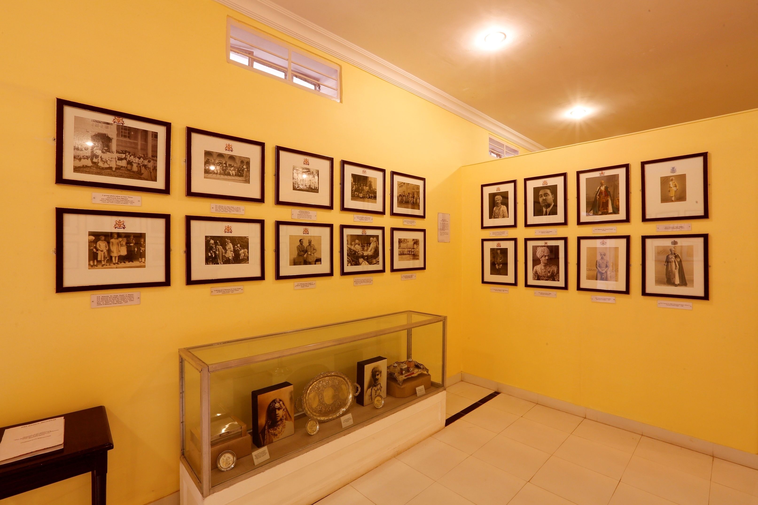 Une galerie où sont exposées des photographies historiques encadrées sur des murs de couleur jaune, une vitrine contenant des objets anciens et une petite table sur laquelle est posé un panneau.