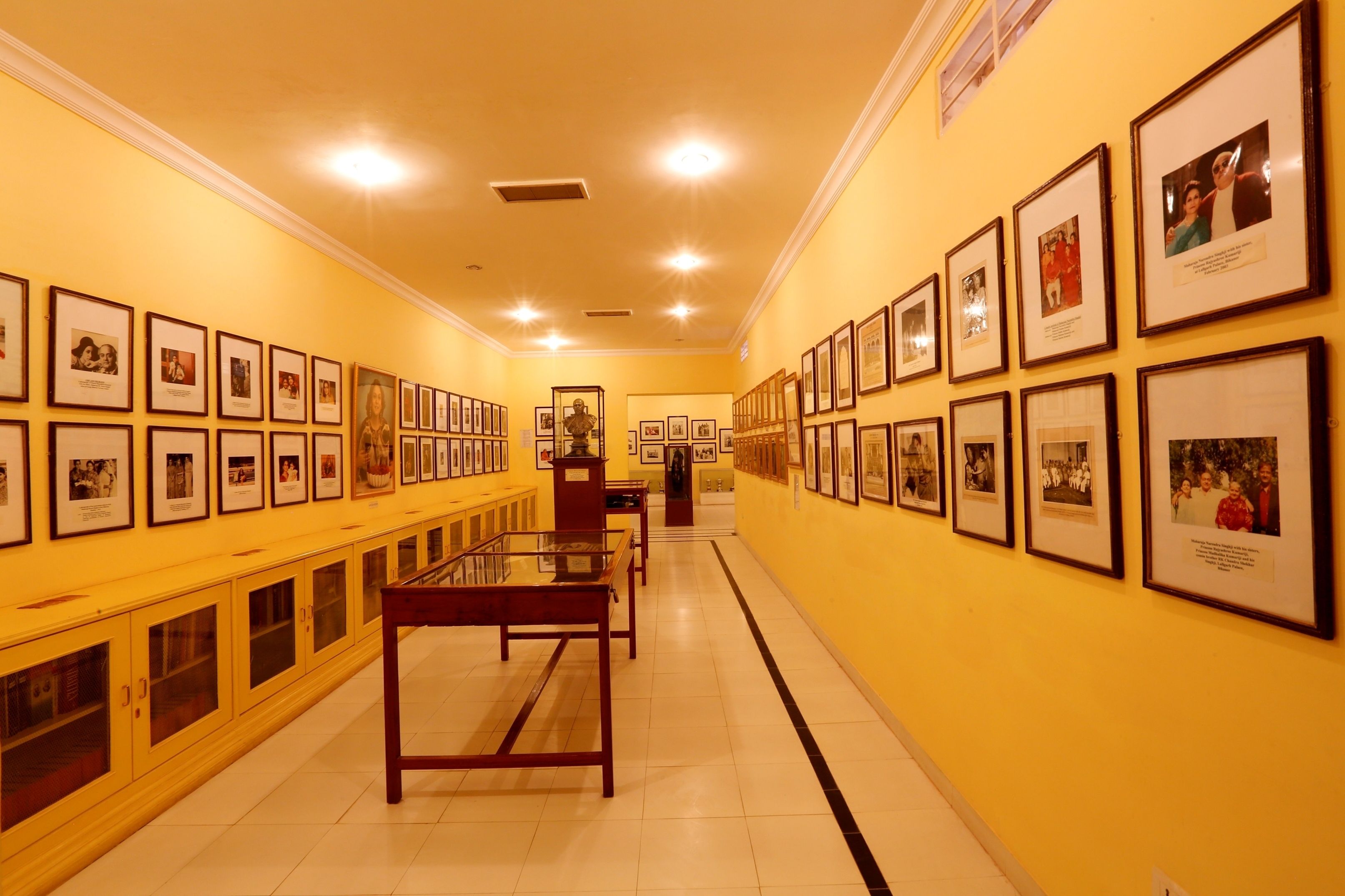 A hallway with framed photographs on the walls, a glass display table, and wooden cabinets along the sides.