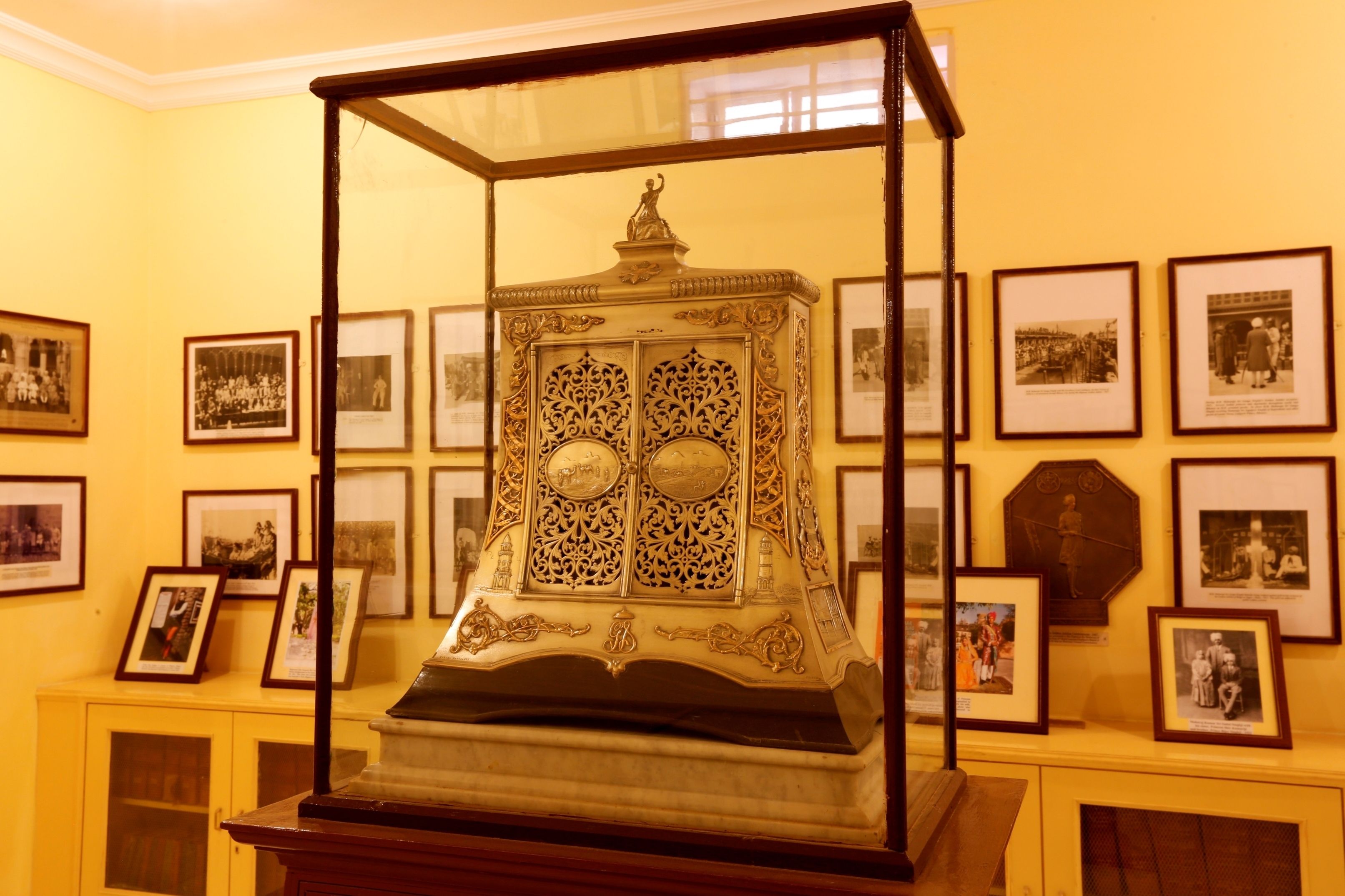 Une vitrine contenant un vase décoratif, entourée de photographies historiques encadrées accrochées au mur.