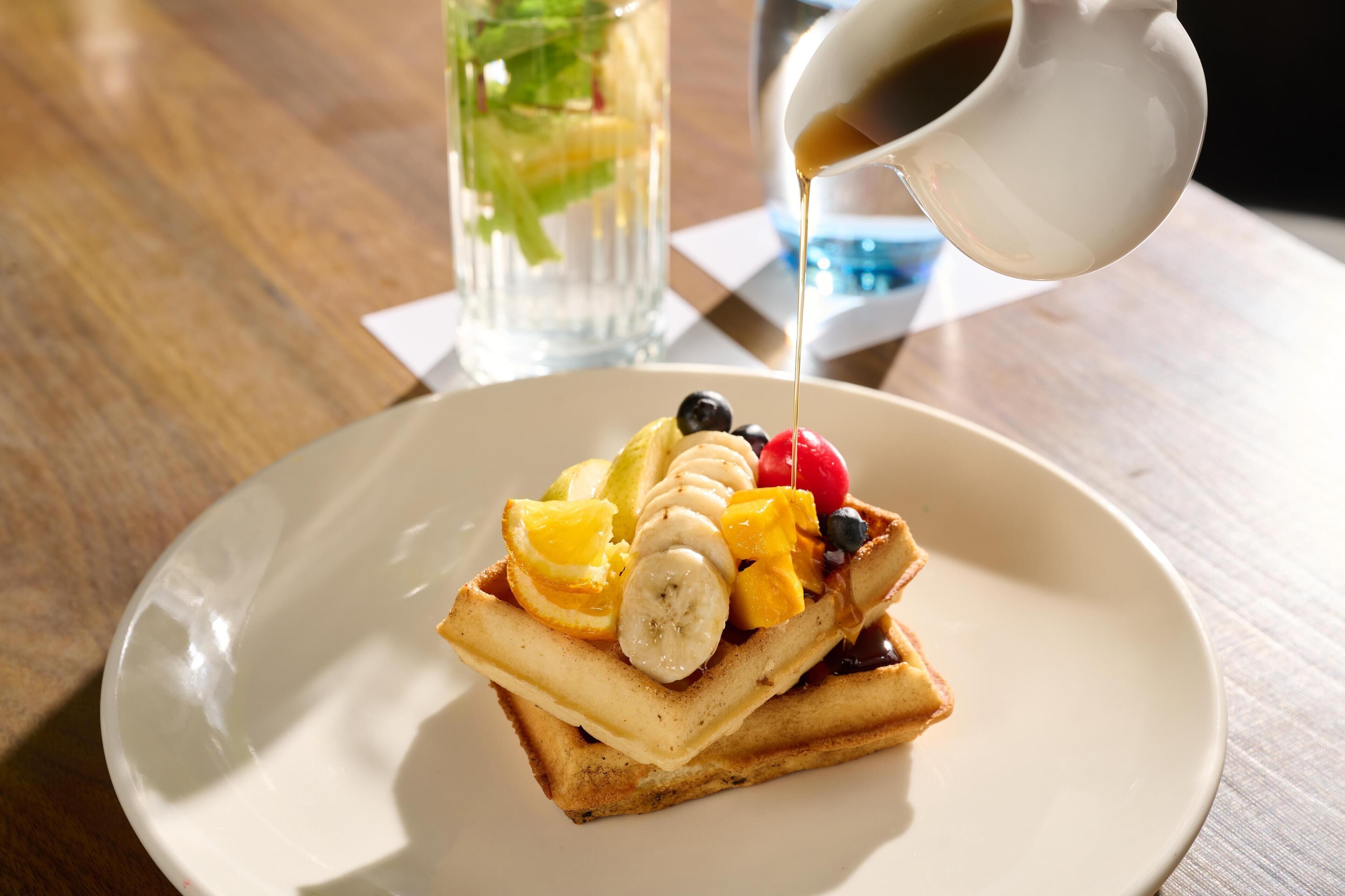 A plate with a stack of pancakes topped with fruit and syrup, a glass of water, and a cup of coffee on a wooden table.