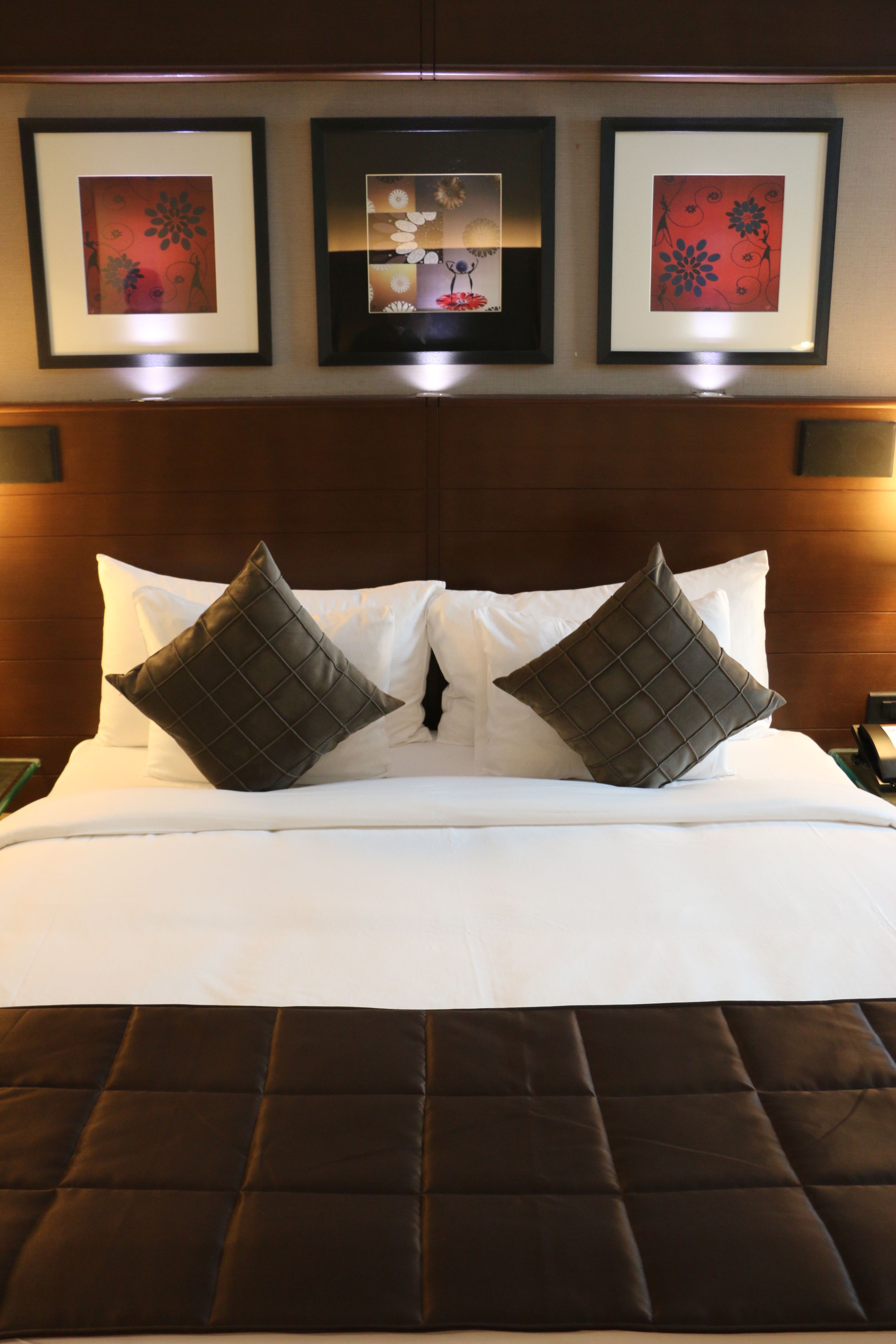 A neatly made bed with two large square pillows, a dark brown quilted headboard, and three framed artworks above the bed.