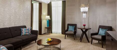 A modern hotel room with a sofa set, two armchairs, a coffee table, and a small side table with a lamp.