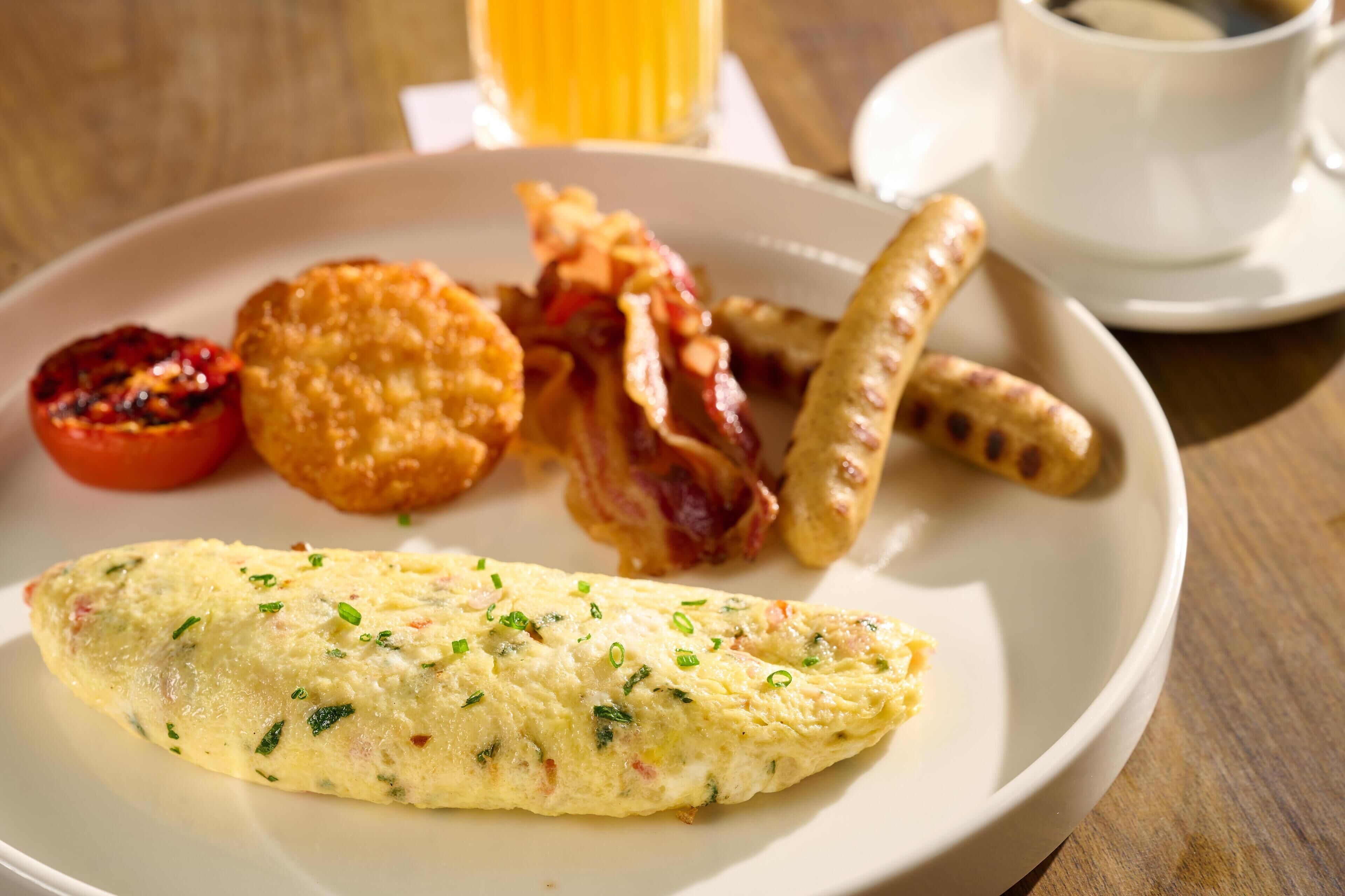 Prato de café da manhã com omelete, bacon, salsichas, tomate e um muffin.
