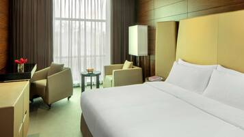 A hotel room with a large bed, two armchairs, a small table with a lamp, and a large window with curtains.