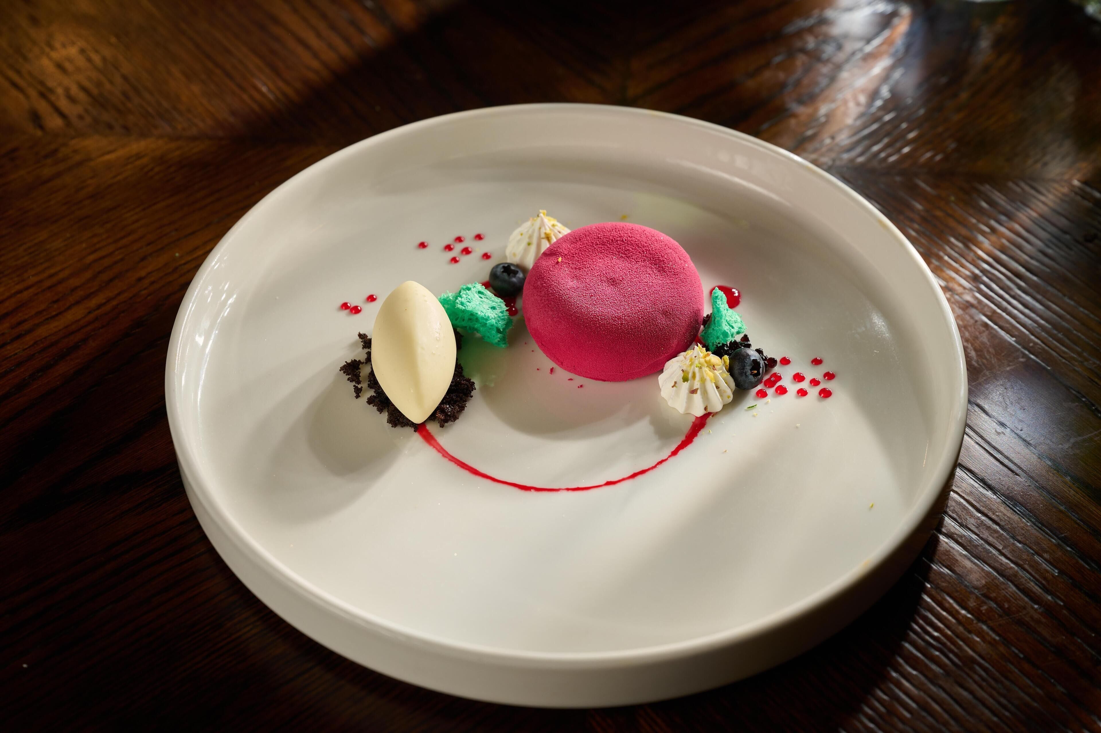 Un dessert servi dans une assiette blanche, agrémenté d’une ligne de sauce rouge, d’une sphère rose et d’autres éléments décoratifs.