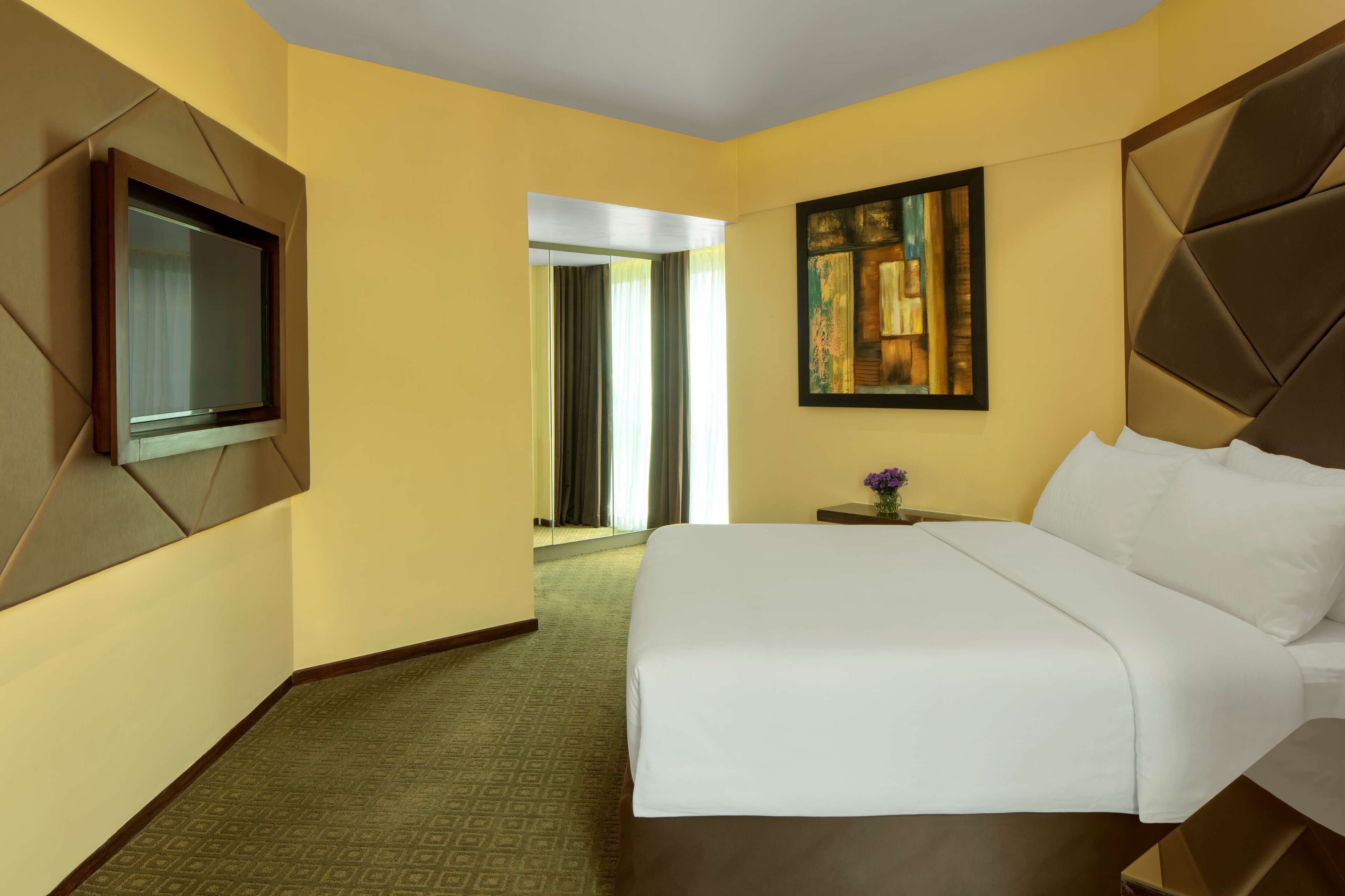 A hotel room with a large bed, a television mounted on the wall, a painting above the bed, and a carpeted floor.