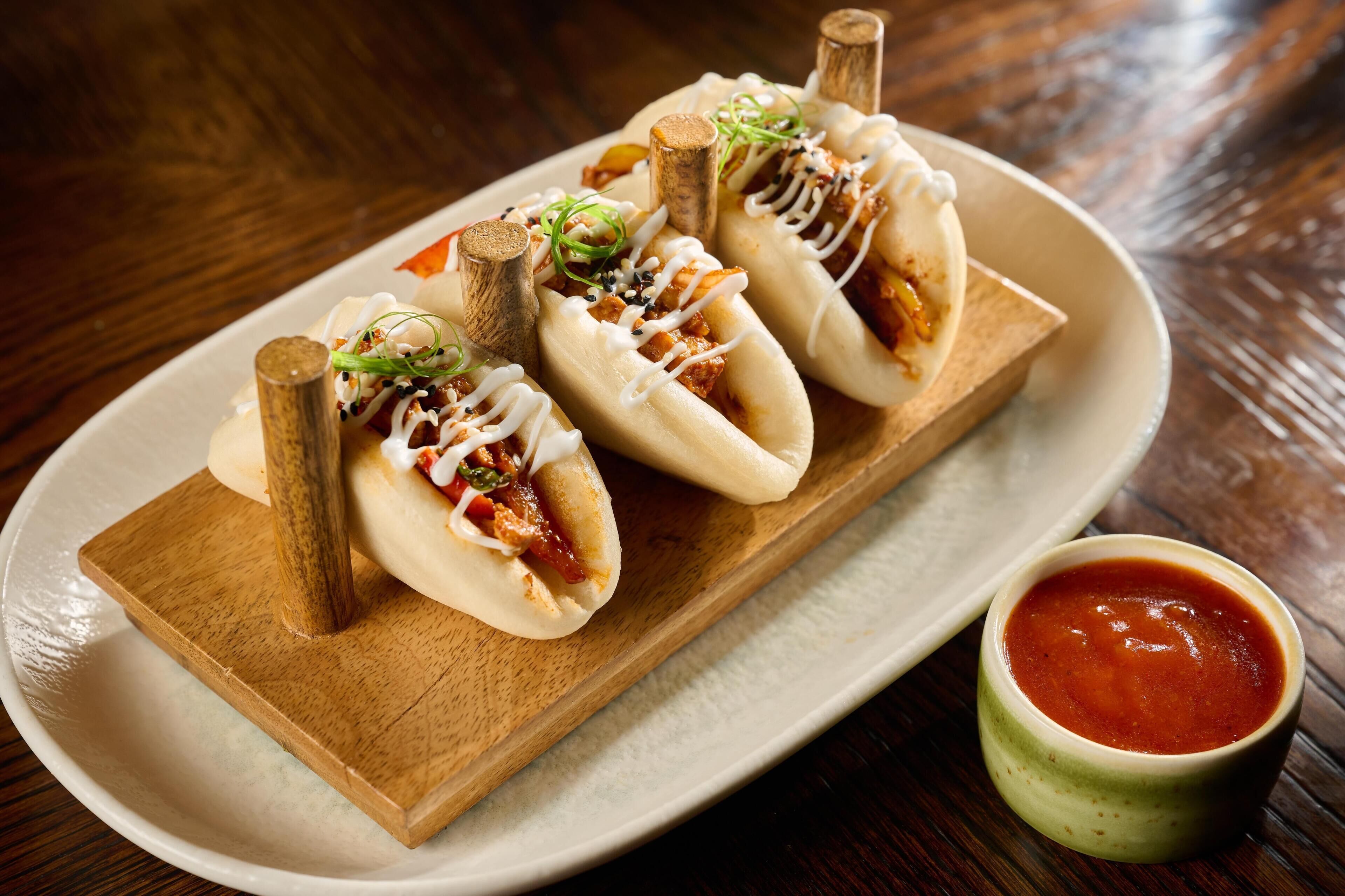 A wooden serving board with three dumplings, each topped with sesame seeds and a drizzle of sauce, accompanied by a small bowl of red sauce.