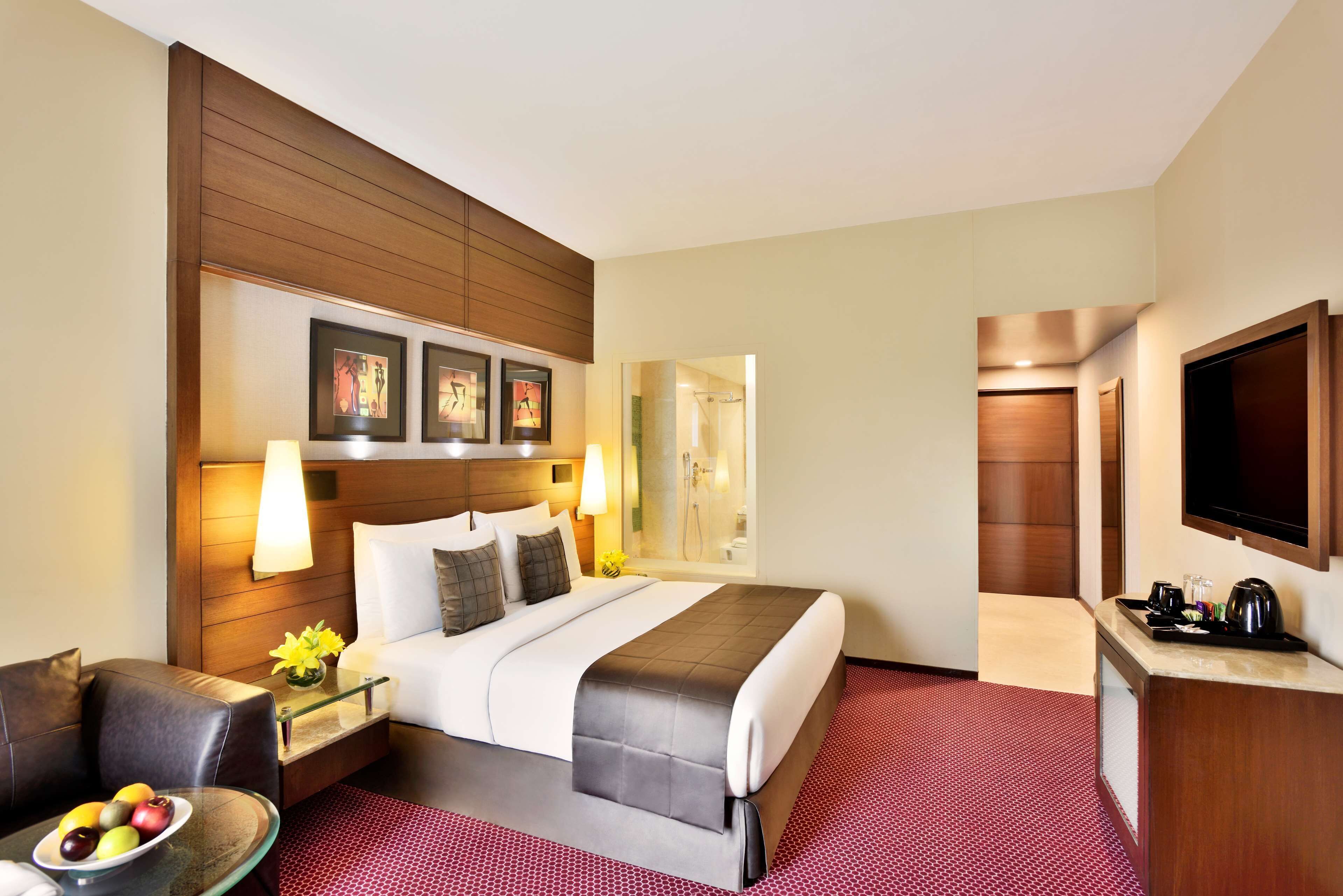 Une chambre d’hôtel avec un grand lit, un bureau avec une lampe, une télévision et un sol recouvert d’une moquette rouge.