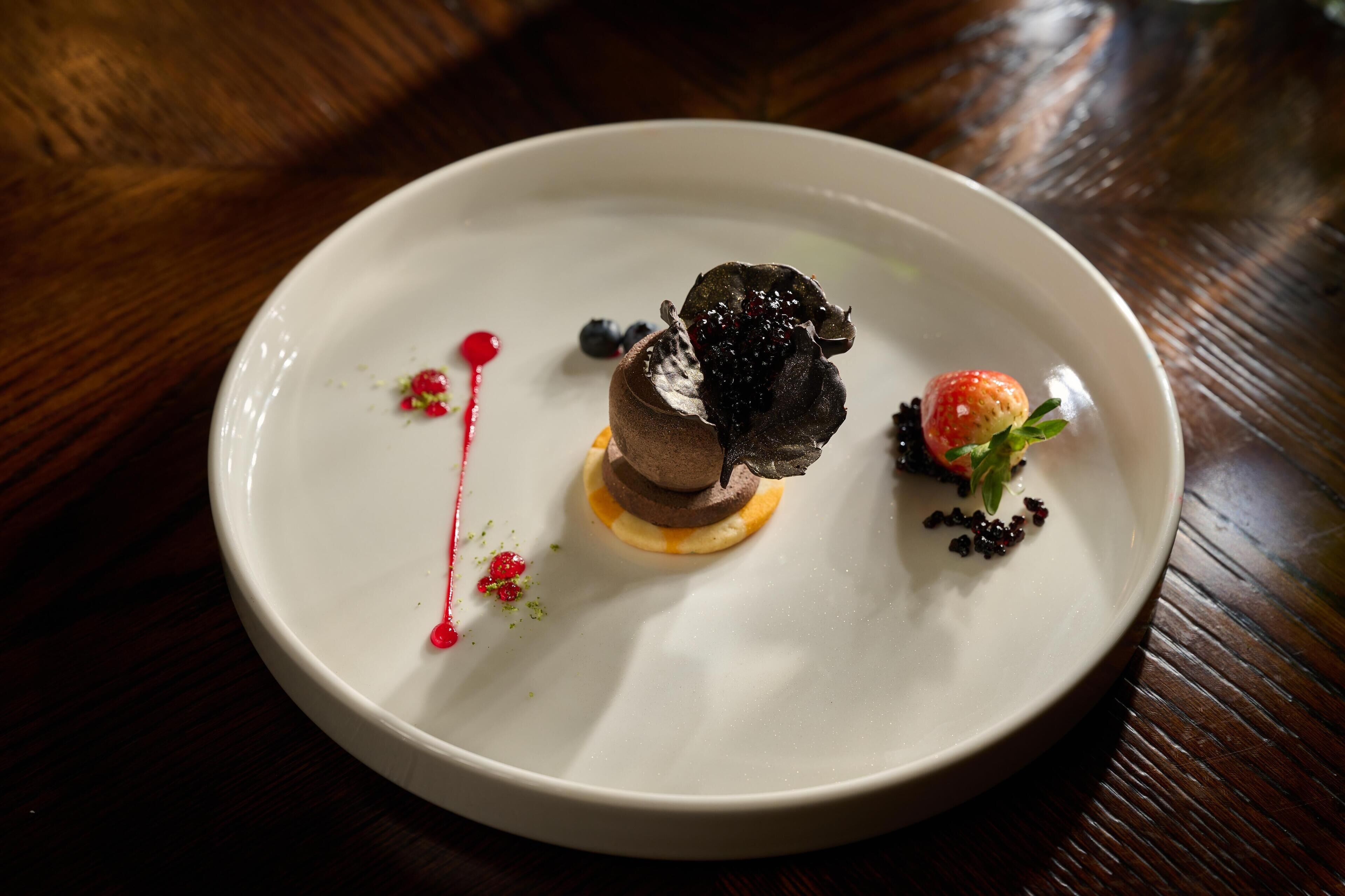 Un piatto bianco con un dolce al centro, guarnito con frutti di bosco e una piccola fragola, su un tavolo di legno.