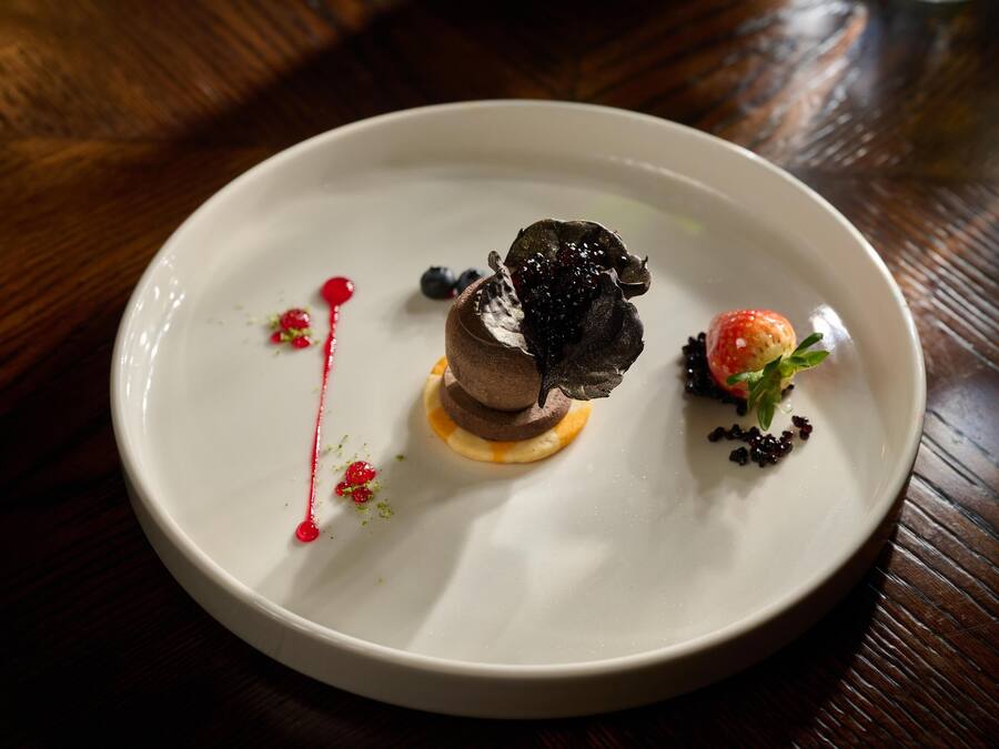 Un piatto bianco con un dolce al centro, guarnito con frutti di bosco e una piccola fragola, su un tavolo di legno.
