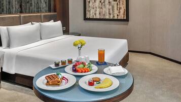 Habitación de hotel con una cama, una mesa de desayuno con platos de comida, un florero con flores y un cuadro en la pared.