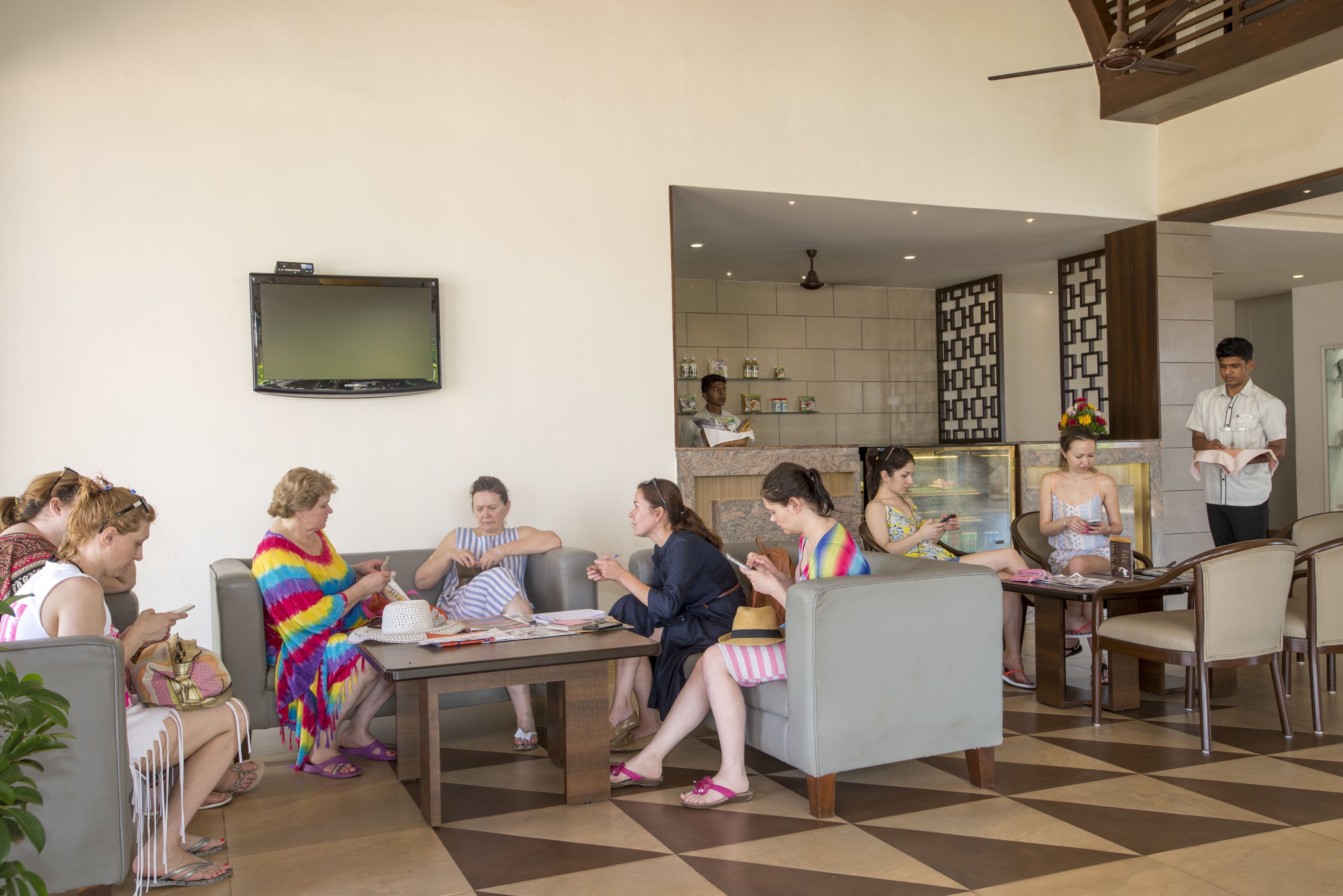 A group of people seated in a modern lobby with a checkered floor, a flat-screen TV mounted on the wall, and a kitchen area in the background.