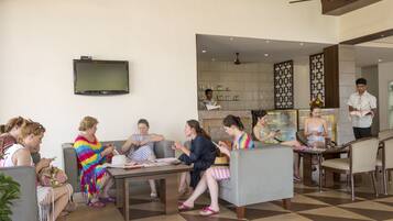 A group of people seated in a modern lobby with a checkered floor, a flat-screen TV mounted on the wall, and a kitchen area in the background.