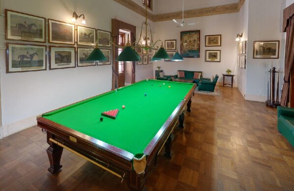 Une salle de billard avec une table de billard, des lampes vertes, des tableaux encadrés aux murs et un plancher en bois.