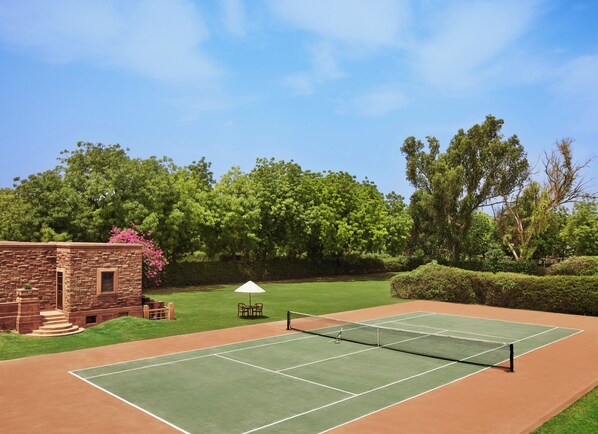 Una cancha de tenis con red, rodeada por un edificio de ladrillo y abundante vegetación.