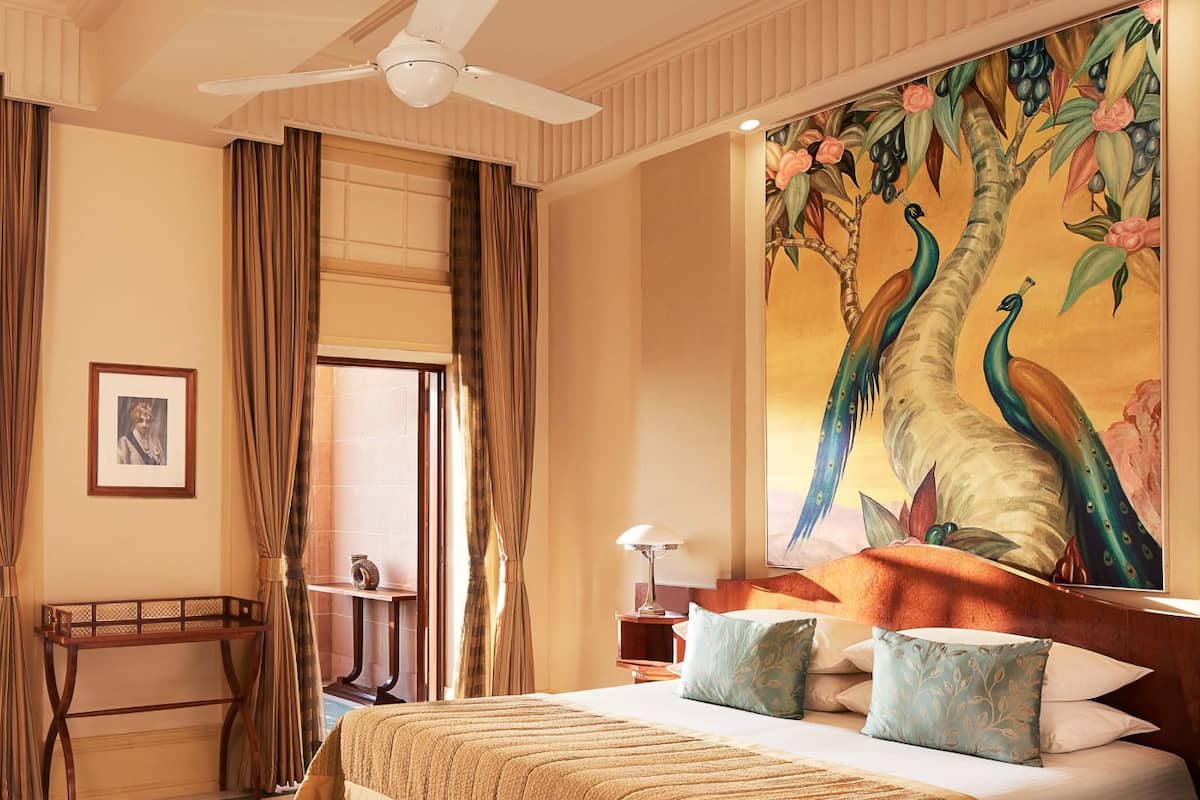 A hotel room with a bed, a ceiling fan, a large painting of a peacock, and a window with curtains.