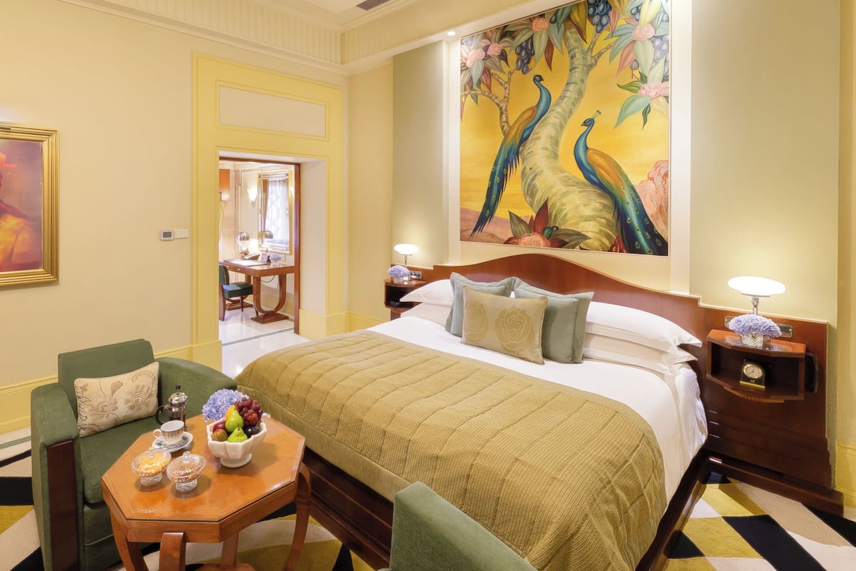 A hotel room with a large bed, a sitting area with a table and chairs, a painting on the wall, and a view of the hallway.