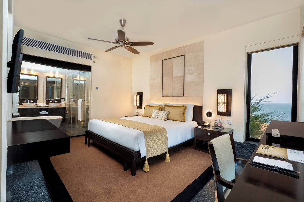 A modern hotel room with a large bed, a desk, a chair, a vanity with a mirror, and a view of the ocean.