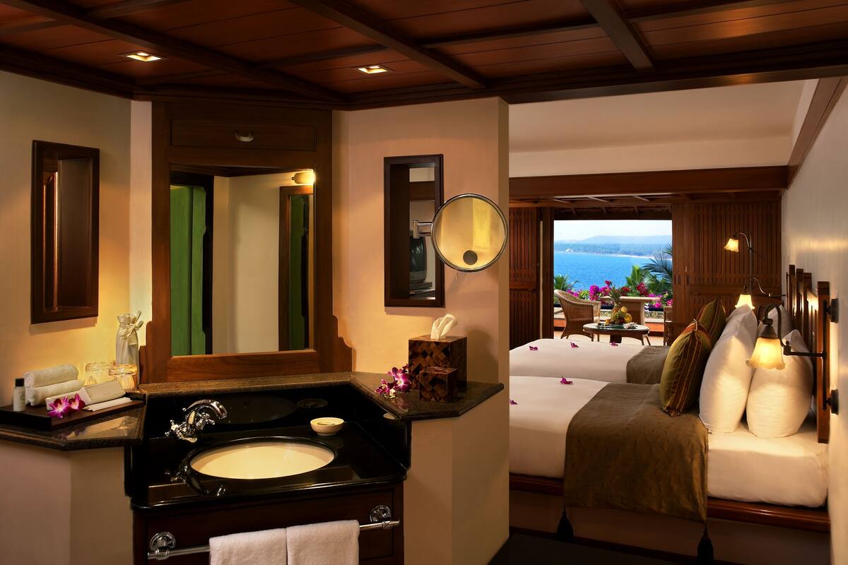 A hotel room with a wooden ceiling, a large window with a view of the sea, a bed with white bedding, and a bathroom with a sink and mirror.