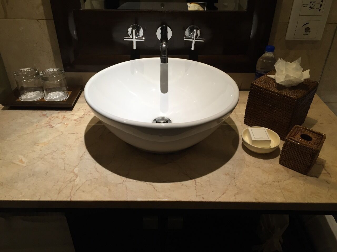 Un lavamanos con lavabo blanco, dos grifos y un espejo encima. Hay dos frascos de vidrio en una bandeja, una caja de pañuelos y un jabonera en la encimera.