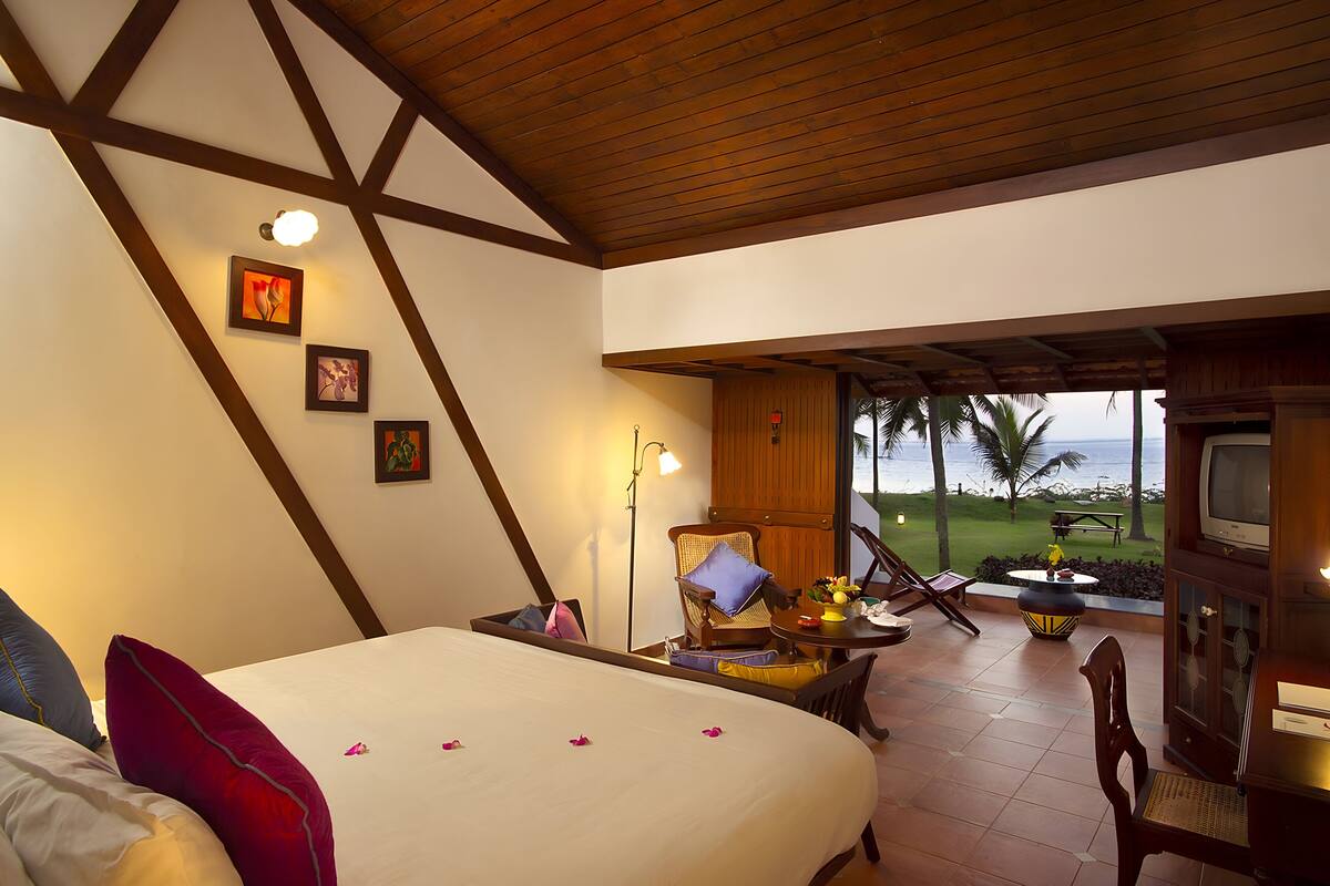 A bedroom with a bed, a chair, a table, and a TV. There is a view of the ocean and palm trees outside.