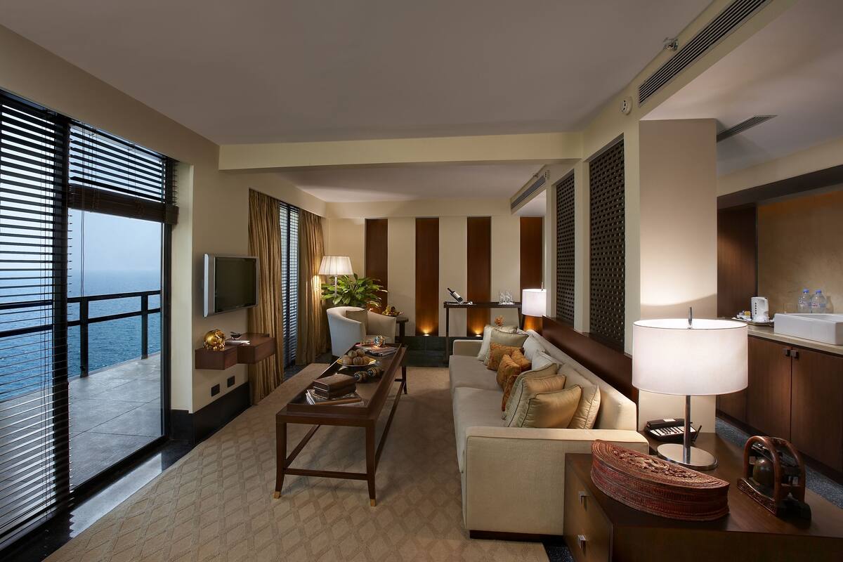 A modern living room with a sofa, coffee table, and TV. There is a large window with blinds, a plant, and a fireplace.