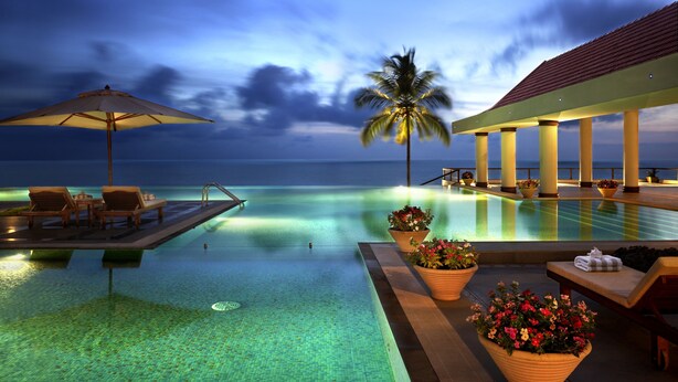 An infinity pool with lounge chairs and umbrellas, a palm tree, and a view of the ocean at dusk. - The Leela Kovalam, a Raviz Hotel (Neyyattinkara)