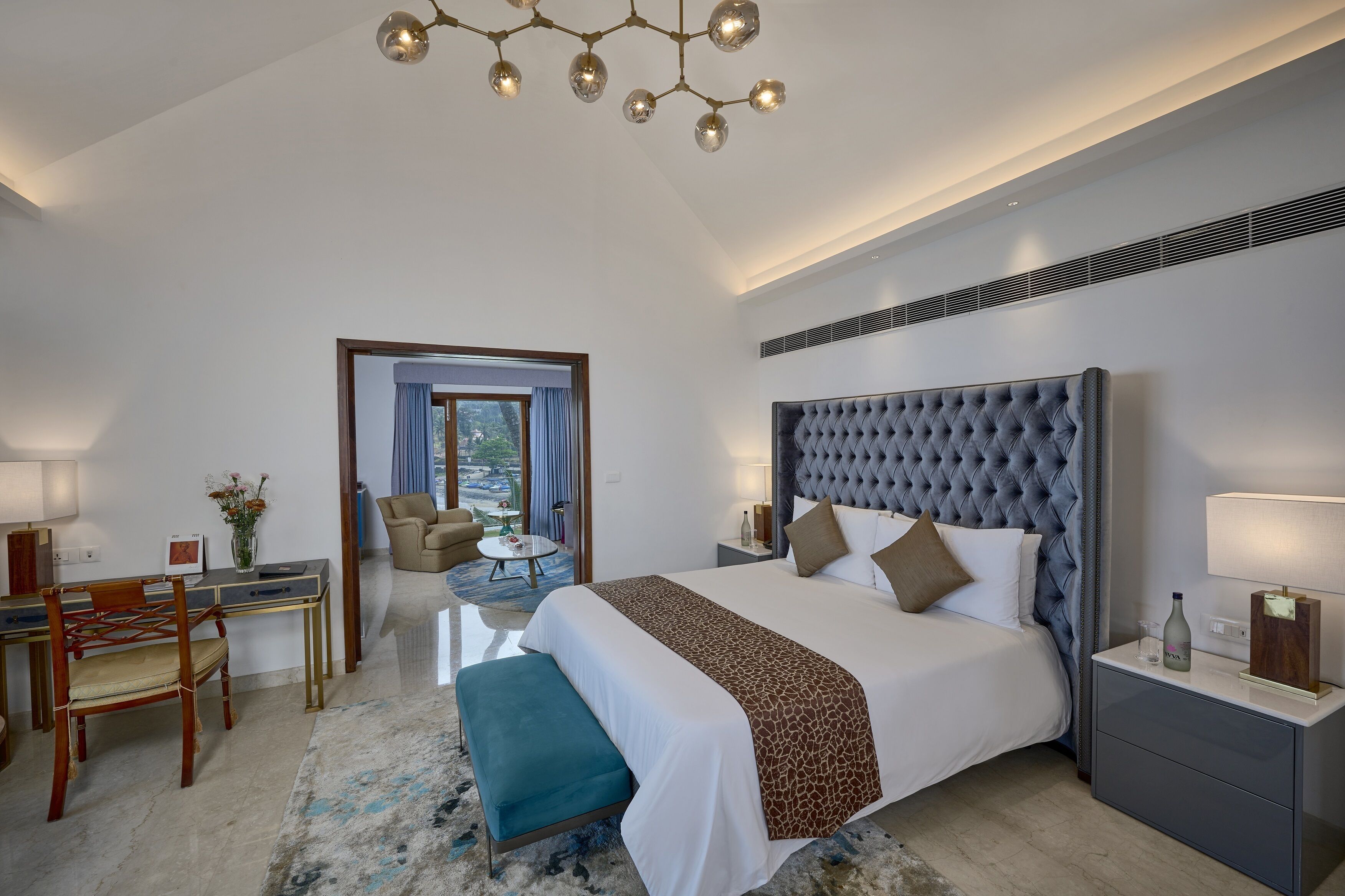 Ein modernes Hotelzimmer mit einem großen Bett, einem Schreibtisch mit Stuhl, einem kleinen Tisch mit Blumenvase und Blick ins Freie.