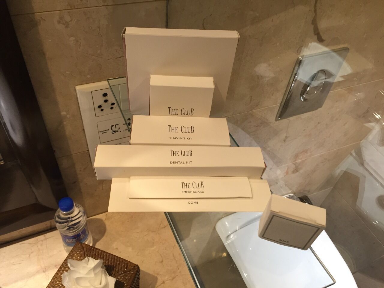 A bathroom with a glass shower enclosure, a white sink, and a mirror. There are several white boxes labeled "The Club" with different items such as a shaving kit, dental kit, emery board, and comb. A bottle of water is also visible on the counter.