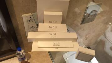 A bathroom with a glass shower enclosure, a white sink, and a mirror. There are several white boxes labeled "The Club" with different items such as a shaving kit, dental kit, emery board, and comb. A bottle of water is also visible on the counter.