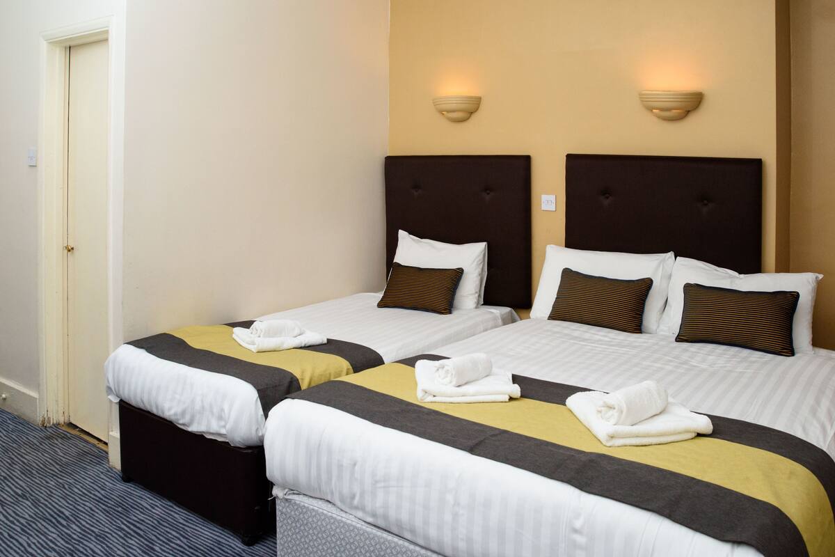 A hotel room with two single beds, each with a headboard, white bedding, and brown pillows. There are wall-mounted light fixtures on the wall above the beds.