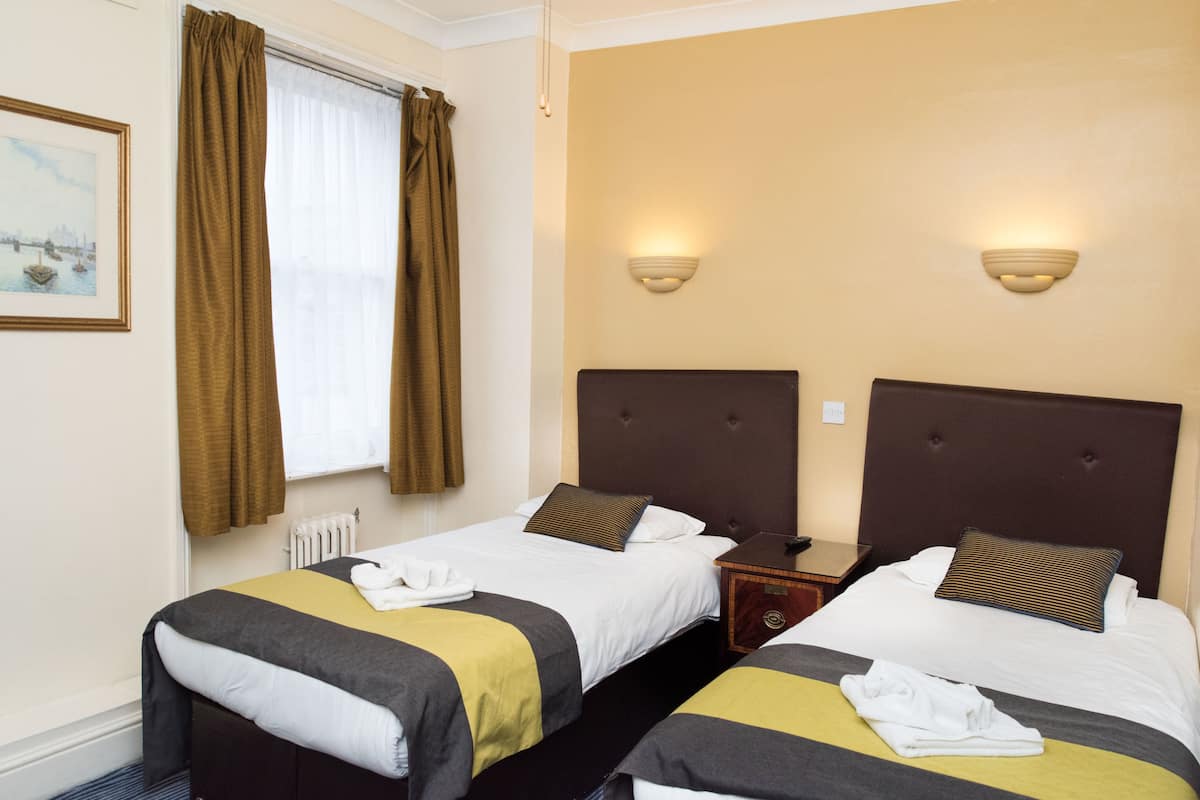 A hotel room with two beds, each with a headboard, a window with curtains, a wall-mounted light fixture, and a framed picture on the wall.
