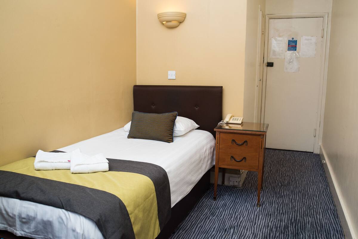 A hotel room with a bed, a bedside table, a wall-mounted light, and a door with a notice.