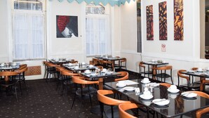 A dining area with tables and chairs, artwork on the walls, and a window with curtains. - Rose Court Hotel (London)