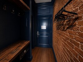 A narrow hallway with a dark blue door, a wooden bench, and a brick wall with a coat rack.