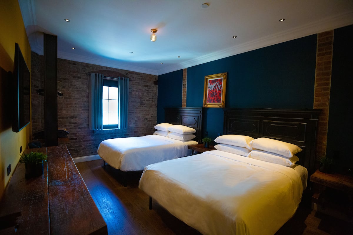 A hotel room with two beds, a wooden bench, a brick wall, a window with blue curtains, and a framed picture on the wall.
