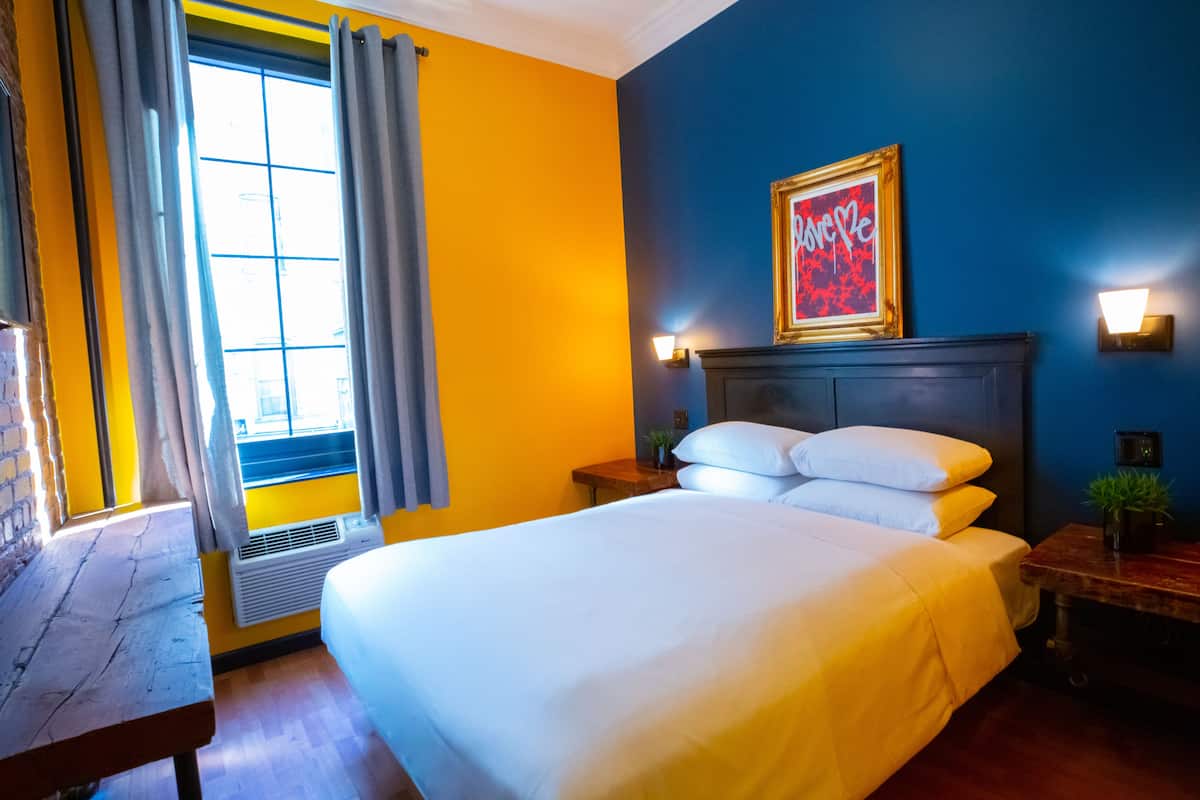 A hotel room with a large bed, a wooden nightstand, a wall-mounted light, and a window with curtains.
