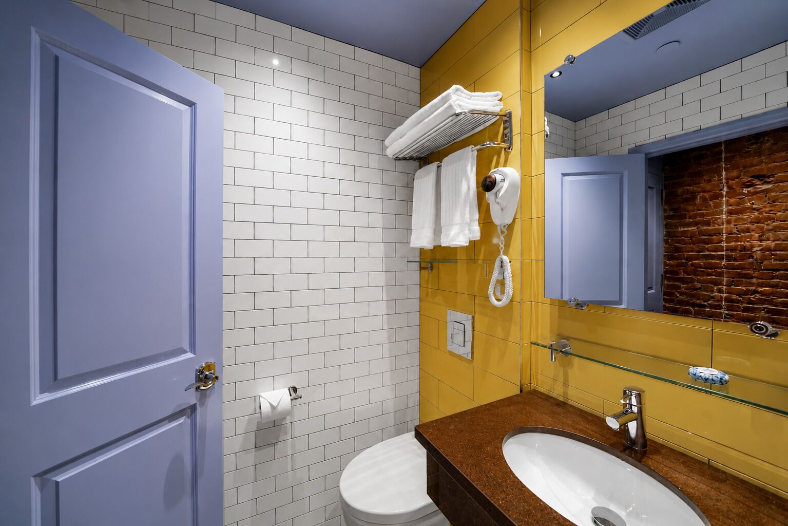 A bathroom with white tiled walls, a yellow accent wall, a glass shower enclosure, a white toilet, and a sink with a brown countertop.