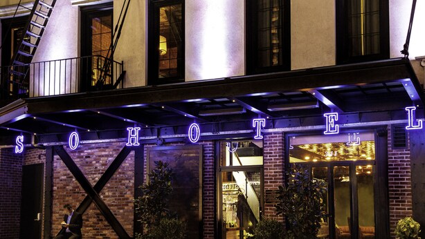 A hotel entrance with a neon sign, brick facade, and outdoor seating area.