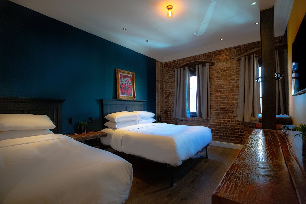 A hotel room with two beds, a brick wall, a wooden bench, and a framed picture on the wall.