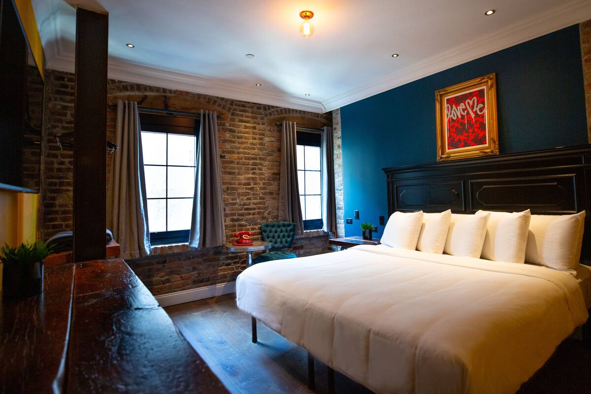 A bedroom with a large bed, a brick wall, a window, and a painting on the wall.