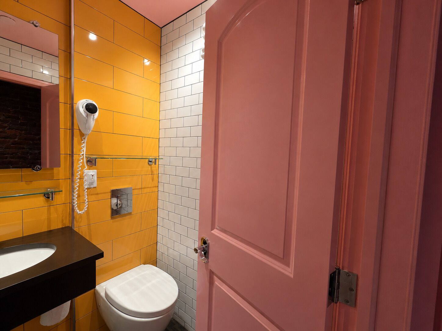 A bathroom with a yellow tiled wall, a white toilet, a black sink, and a pink door.