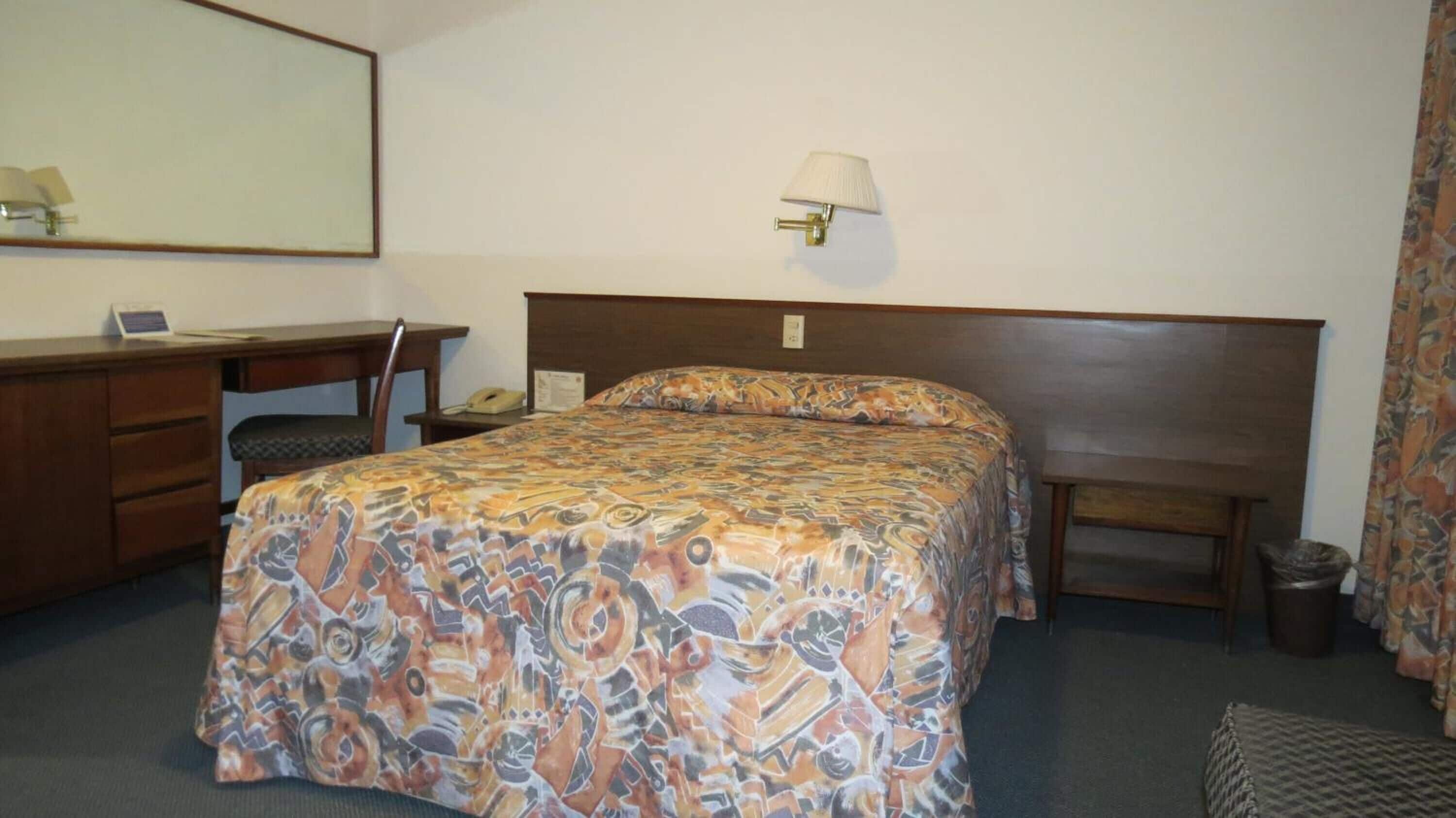 Een hotelkamer met een bed, een bureau, een stoel, een lamp en een prullenbak.