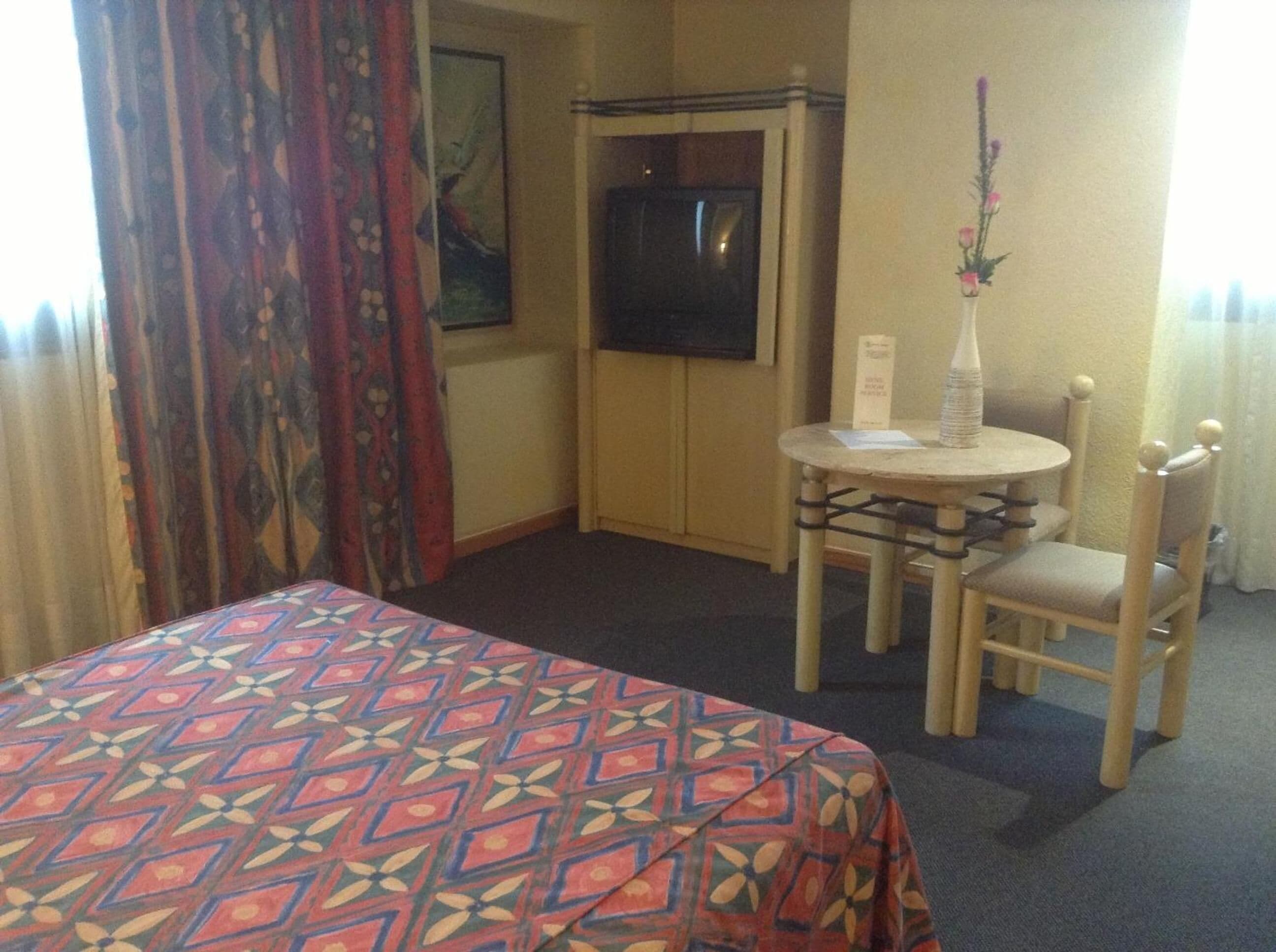 Quarto de hotel com uma cama, uma mesinha com um vaso e uma cadeira.